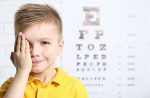 A young boy in a yellow shirt is covering his right eye with his palm, undergoing a vision acuity test at the optician's clinic.