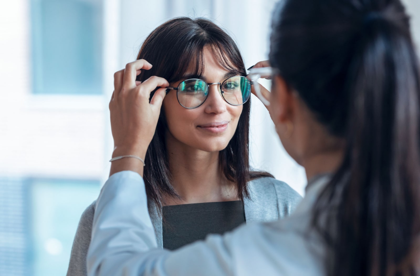 An optician places glasses on a young woman face, helping her fit the glasses for the other woman