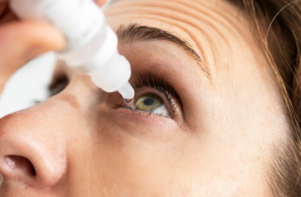 A close-up image of a woman applying eye drops to her left eye.