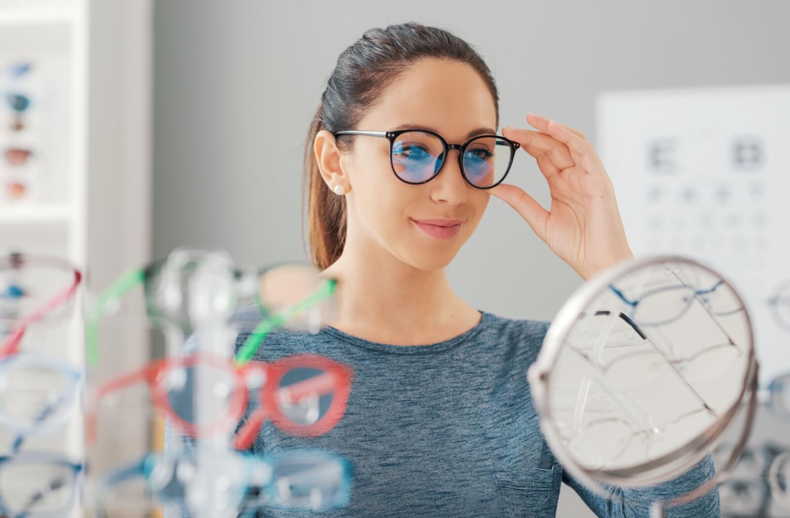 A woman with an oval face smiling in an optometry clinic while trying on a new pair of eyeglass frames.