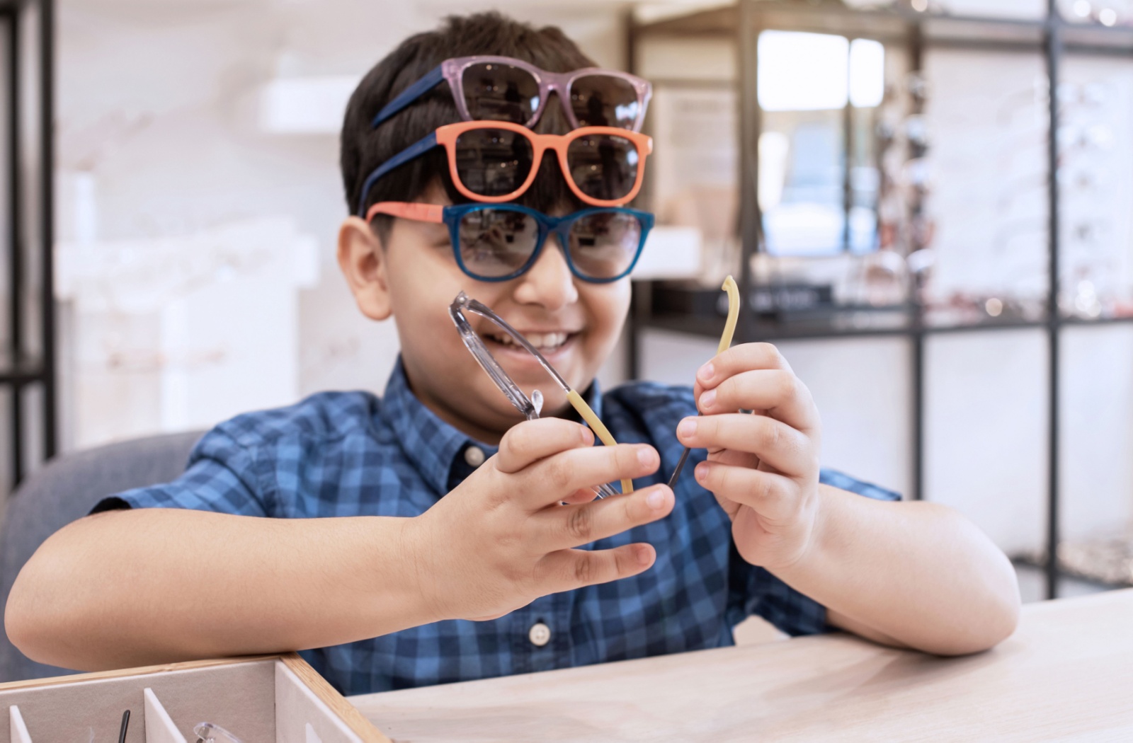 A smiling child wears three pairs of eyeglasses on their head while holding a fourth pair at the optometrist.