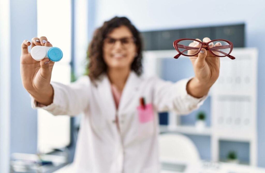 An optometrist wearing a white coat holds up a set of contacts in one hand and a pair of glasses in the other.