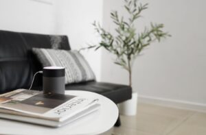 A humidifier releasing mist on a white table beside a magazine in a minimalist living room with a black sofa and a potted plant in the background.