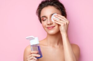 Against a pink background, a young adult smiles while cleaning their eyelids with a mild, hypoallergenic eyelid cleanser.